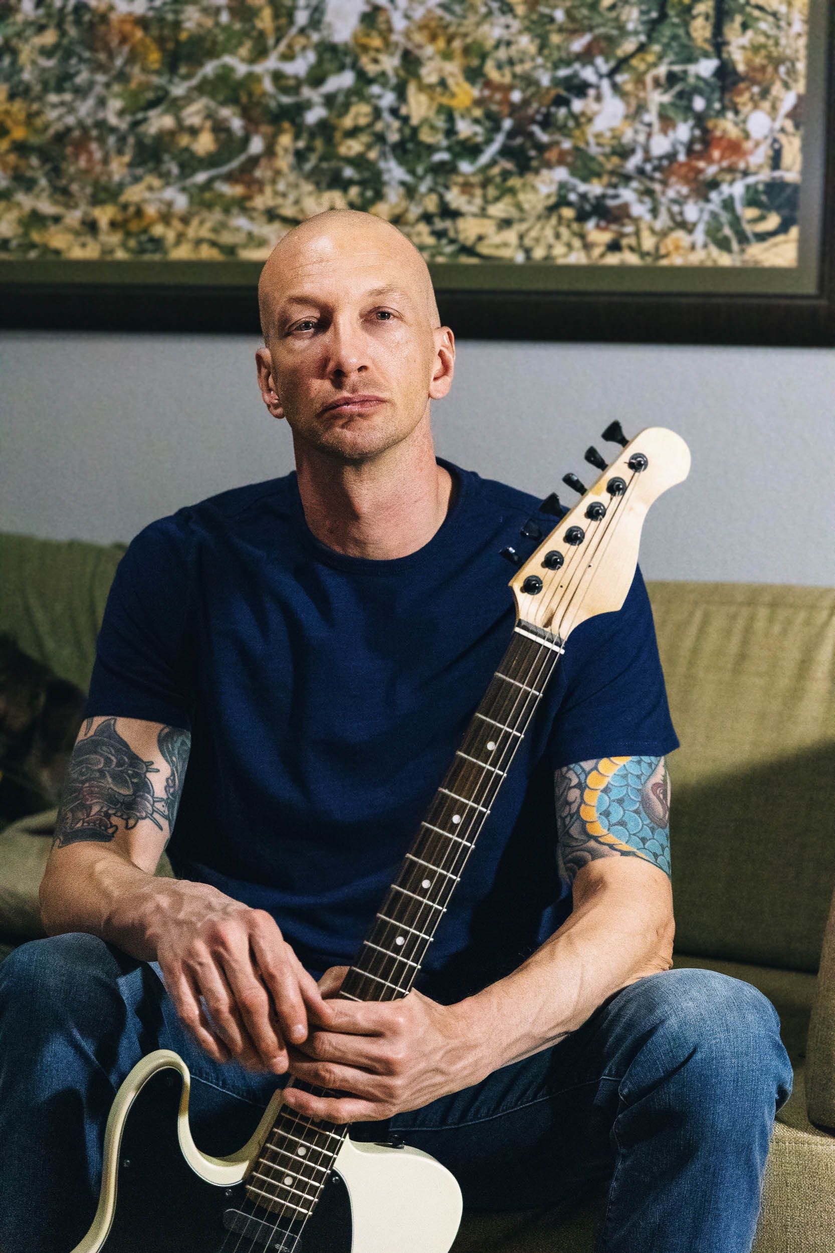 man sitting on a couch in a blue t shirt and jeans holding a white electric guitar.