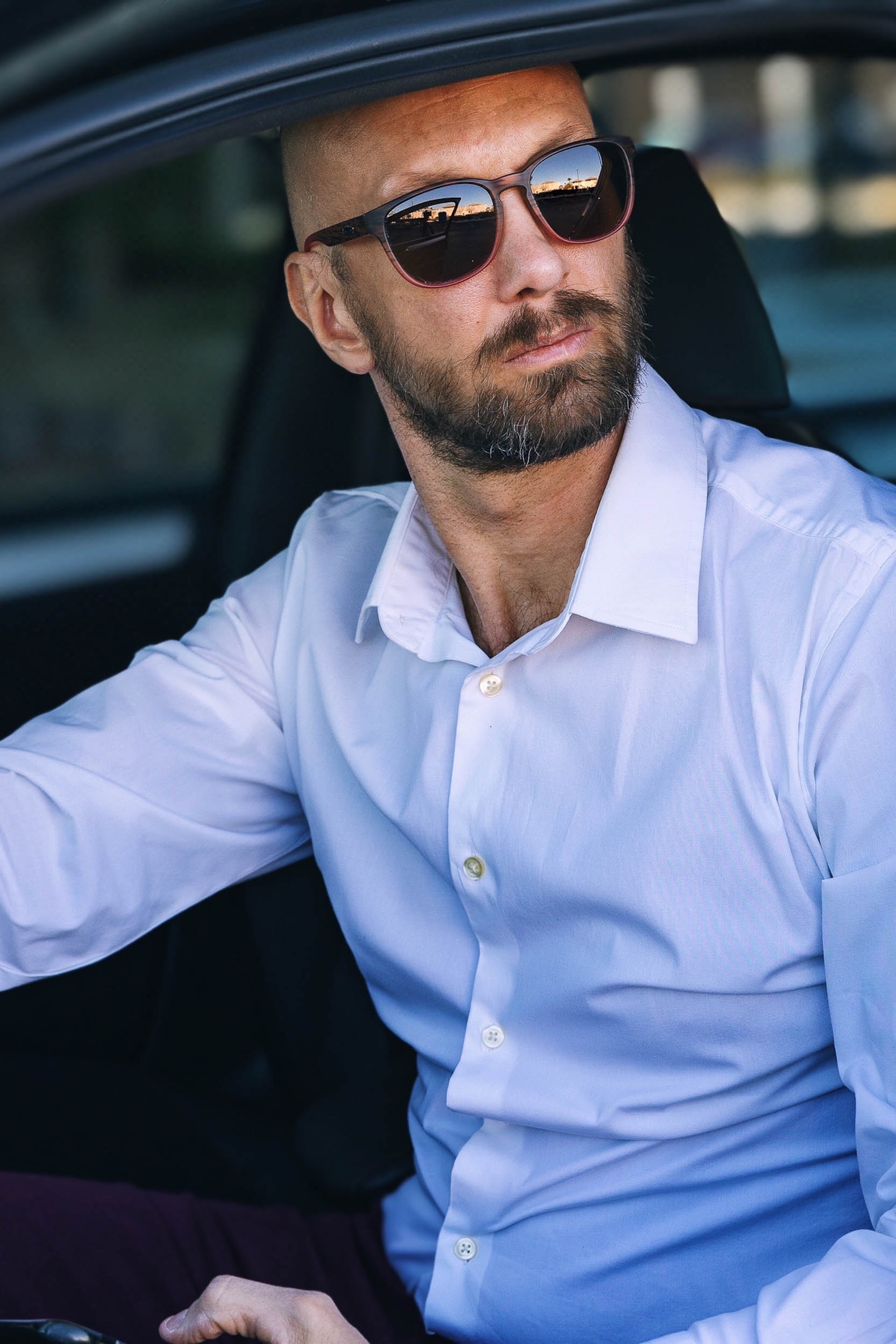 a man in a white dress shirt and sunglasses sitting in a car holding his phone