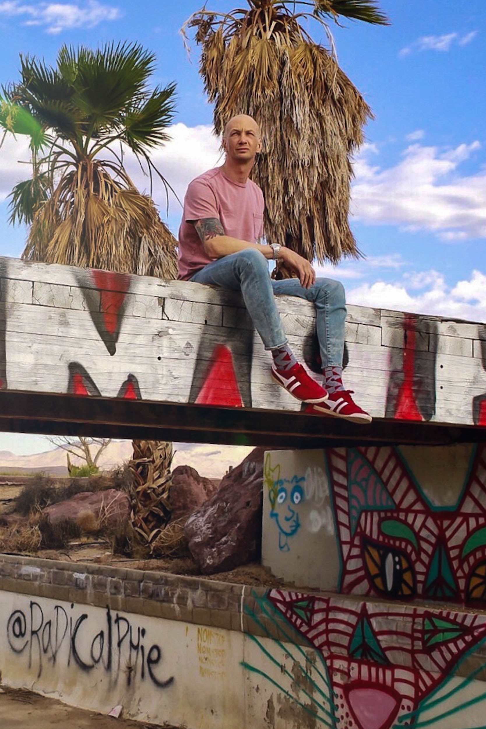 a guy in a pink t shirt sitting own a bridge thats covered in graffiti.