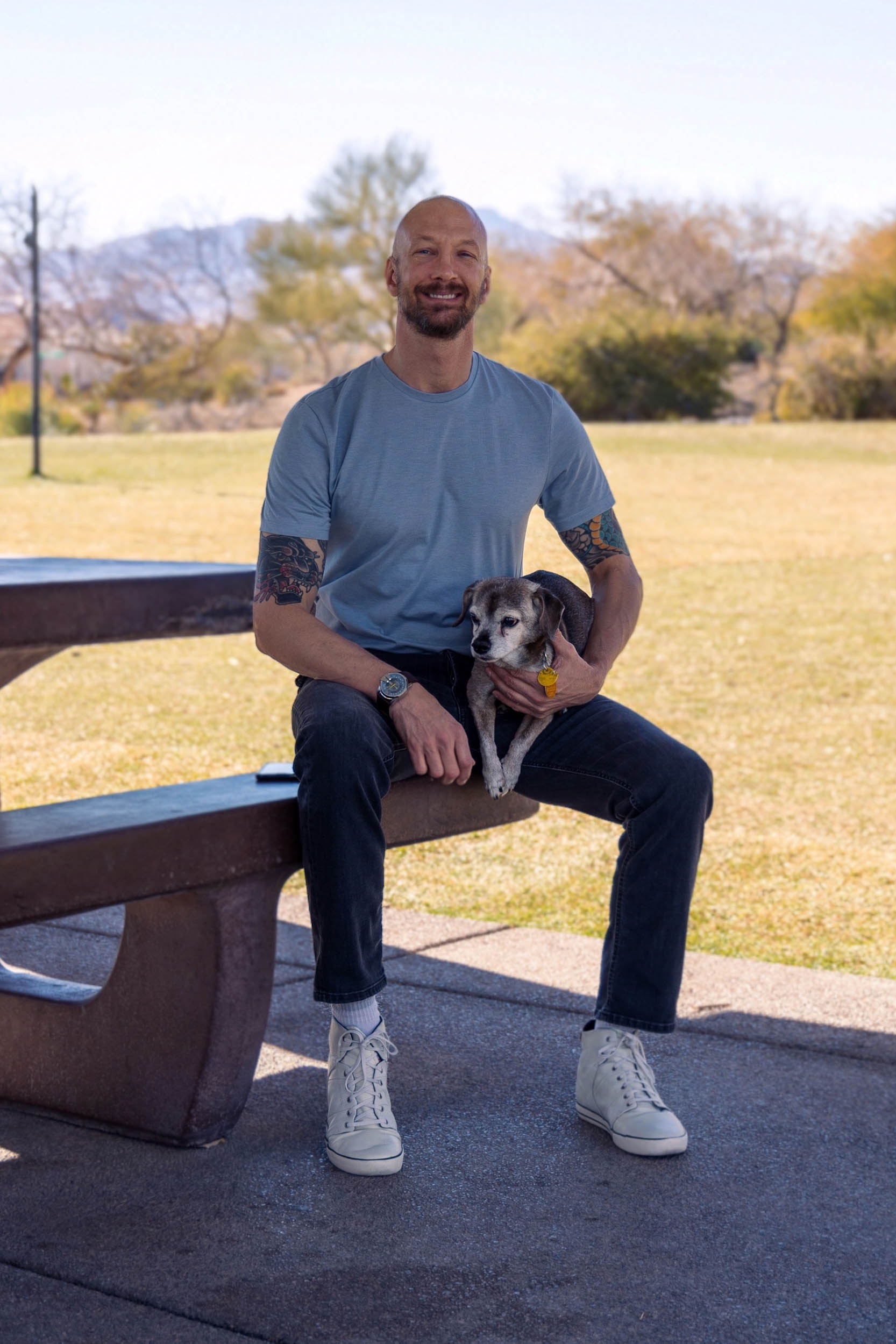 a smiling man in a casual outfit sitting at a stone picnic table holding his dog.