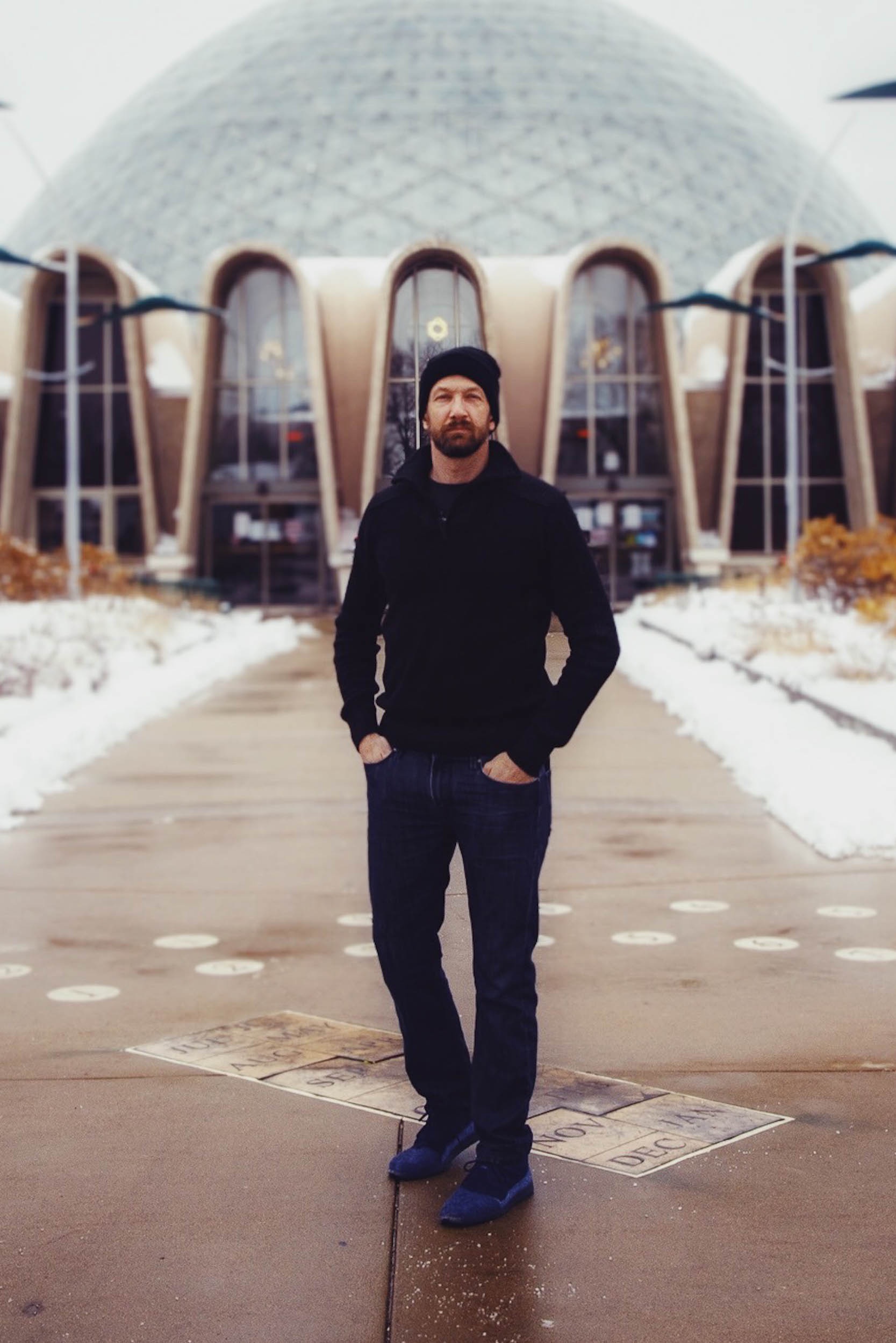 man in a black sweater and hat standing outside a geodesic dome.