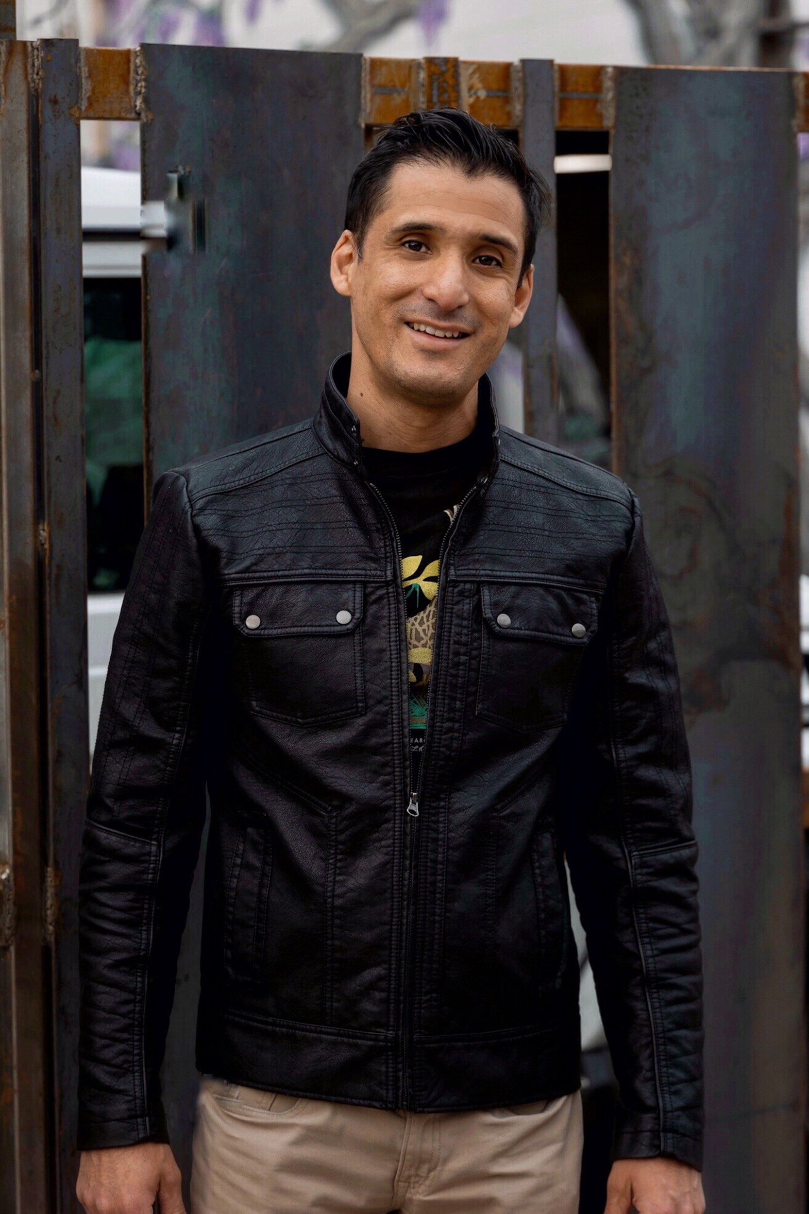 a smiling man in a black jacket next to a rusty fence.