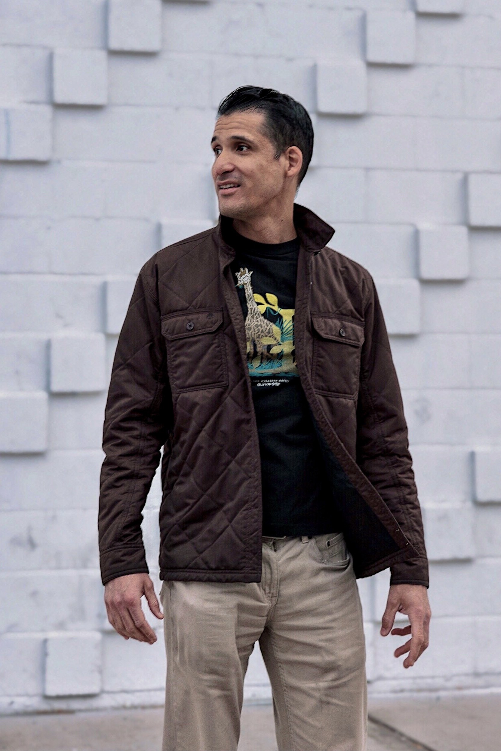 a smiling man in quilted brown jacket standing in front of a white brick wall.