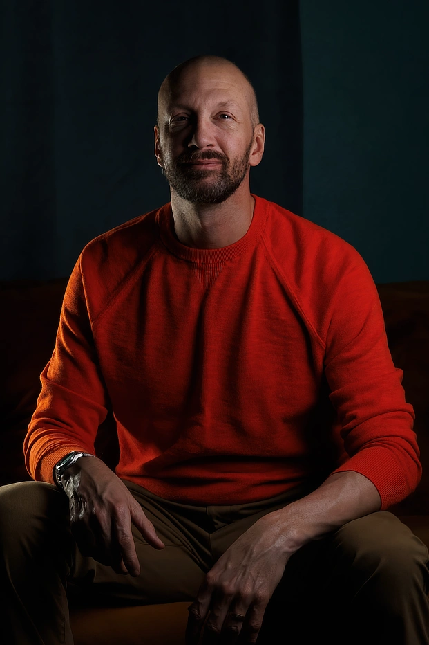 A self portrait of Las Vegas photographer DBA flashqbe in an orange sweater, seated in moody teal lighting, conveying a fun, confidence and creativity with an inviting smile.