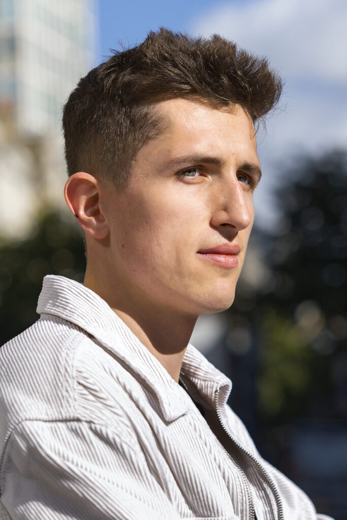 A young man wearing a white corduroy jacket looking into the distance during an outdoor portrait session in natural sunlight.