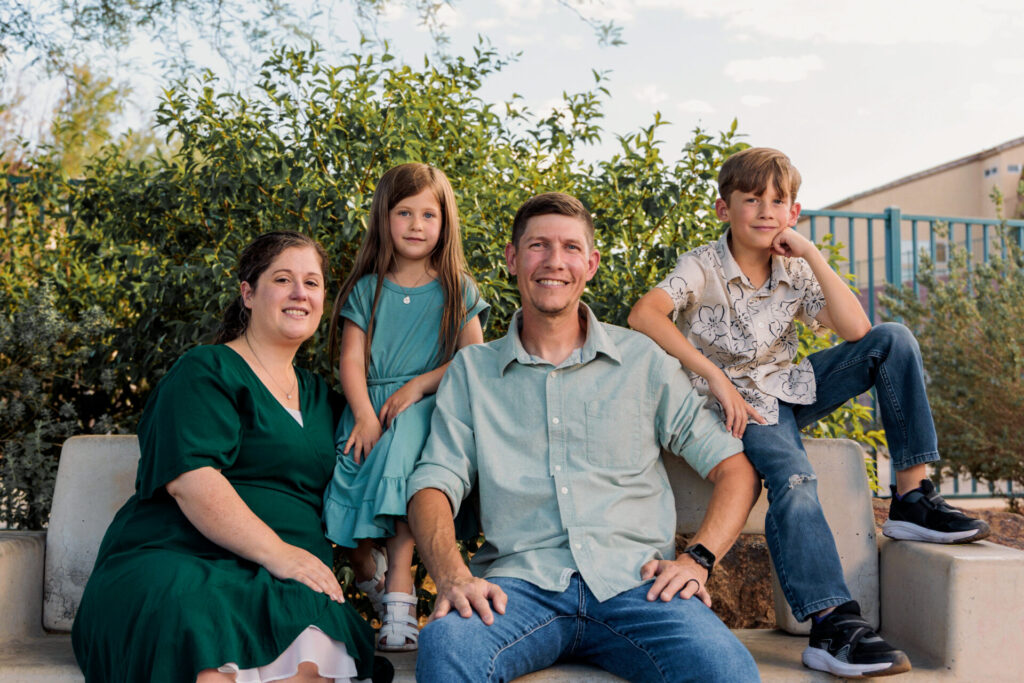 A family of four sits close together on a concrete park bench, smiling together for a professional portrait.