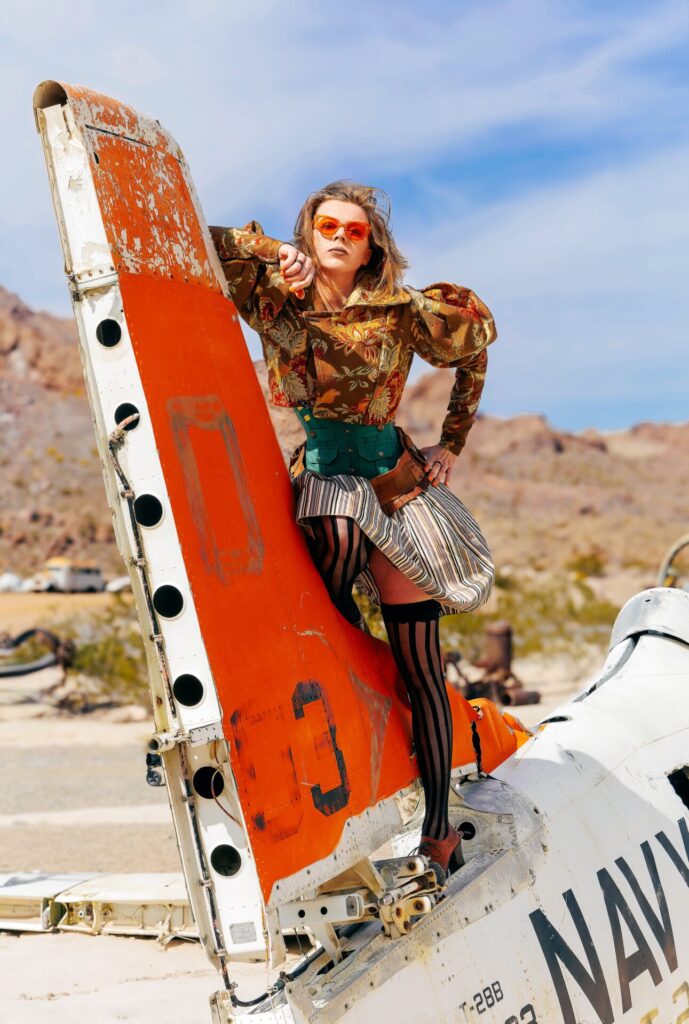 A woman in an edgy brown & orange steampunk outfit is posed on top of a crashed airplane in Nelson’s ghost town.