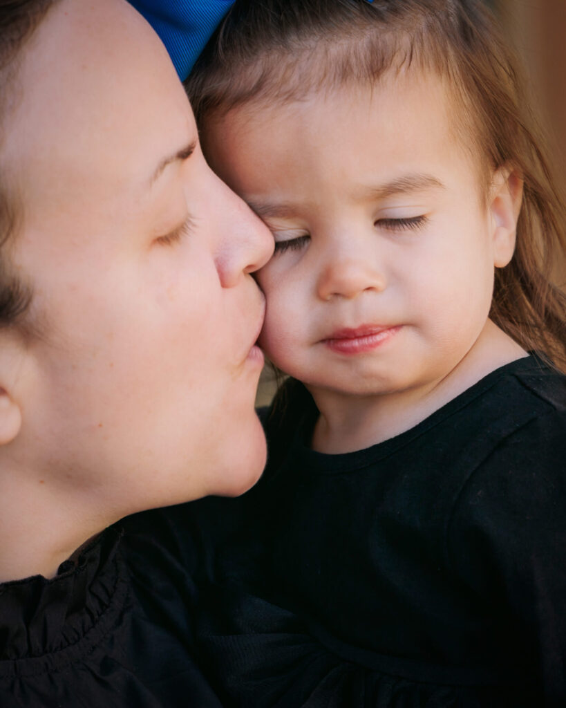 Mother gently kissing her young daughter’s cheek as the child closes her eyes, both wearing black clothing and a soft expression.