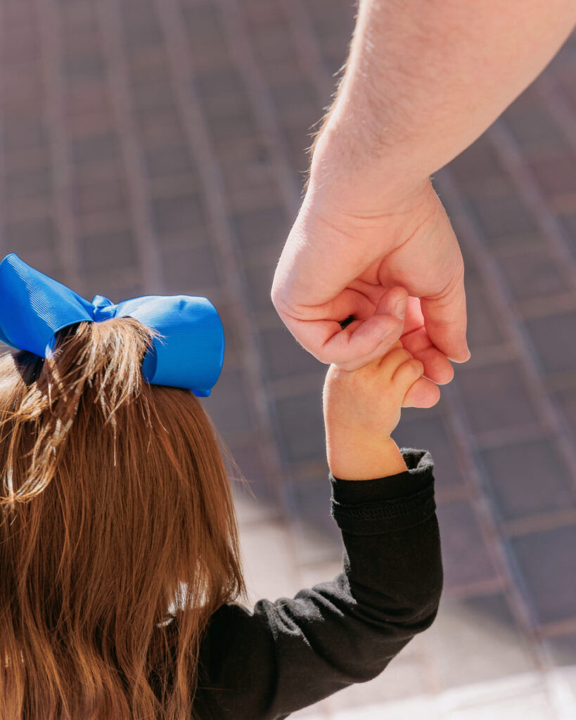 Close up of a young child holding an adult’s hand while walking outdoors, wearing a bright blue bow and black shirt