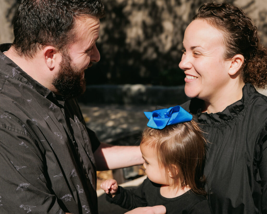 Parents smiling and holding their young daughter with a blue bow during a sunny outdoor family photo session