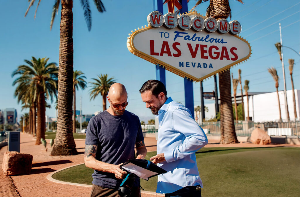 A photographer is showing his client photos on the back of his camera. They are doing a photoshoot in front of the Welcome To Las Vegas sign.