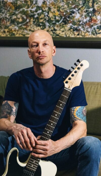 man sitting on a couch in a blue t shirt and jeans holding a white electric guitar.