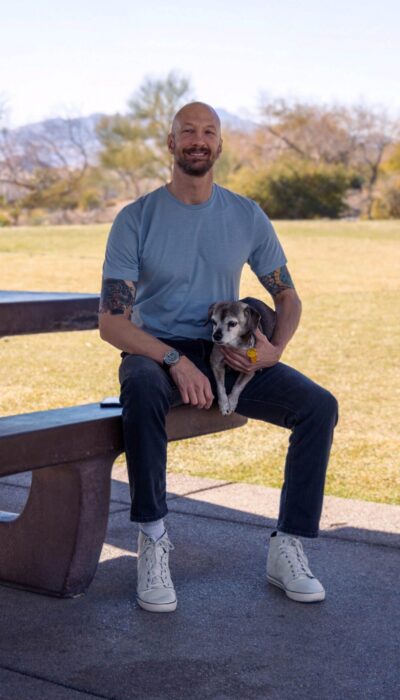 a smiling man in a casual outfit sitting at a stone picnic table holding his dog.