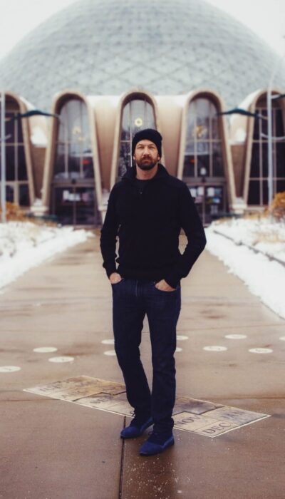 man in a black sweater and hat standing outside a geodesic dome.